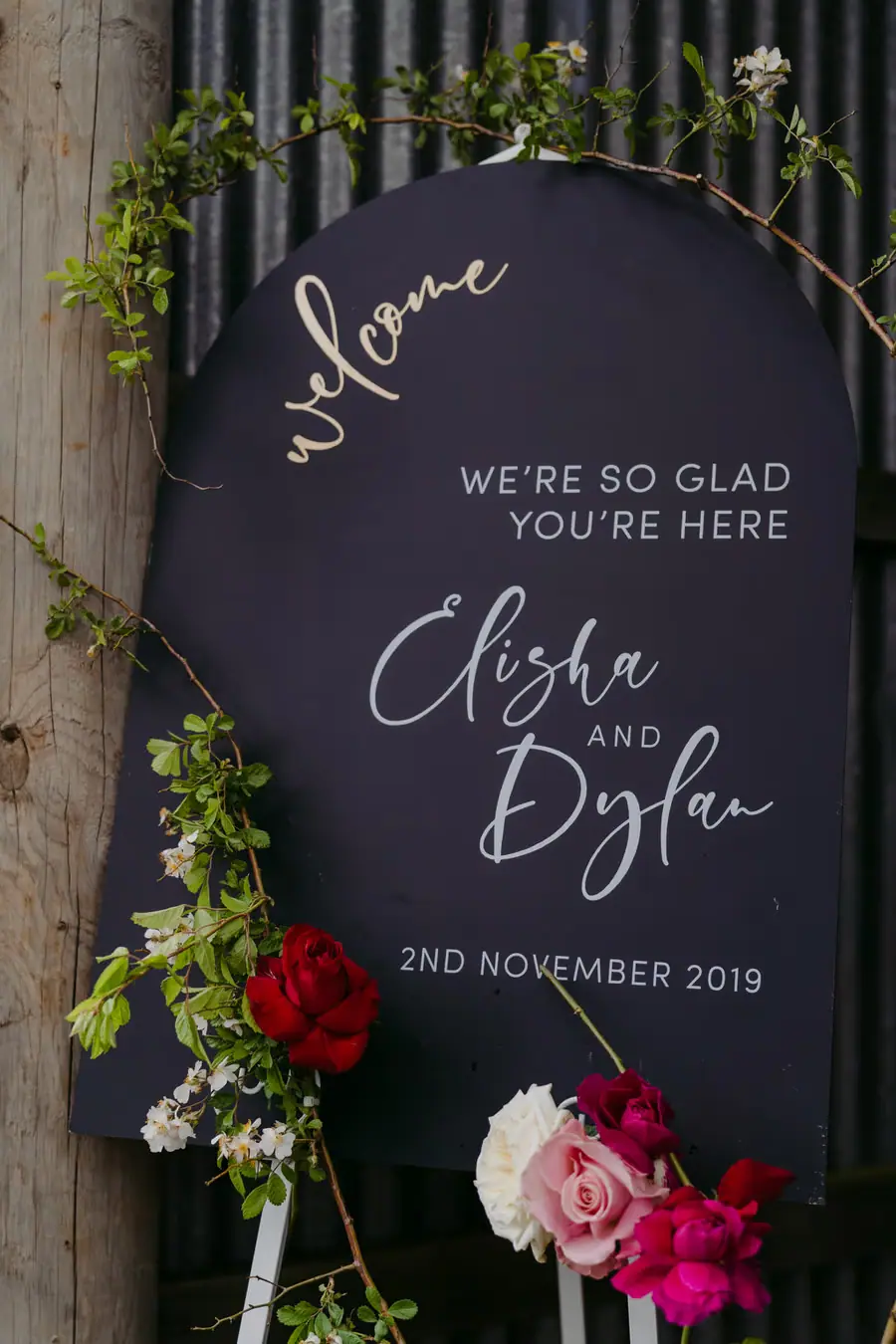 Black wedding welcome sign framed by colourful flowers and greenery.