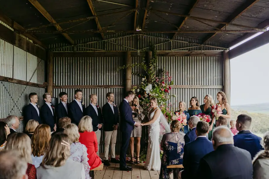 Wedding party standing among guests beneath the metal roof of a rustic ceremony room.