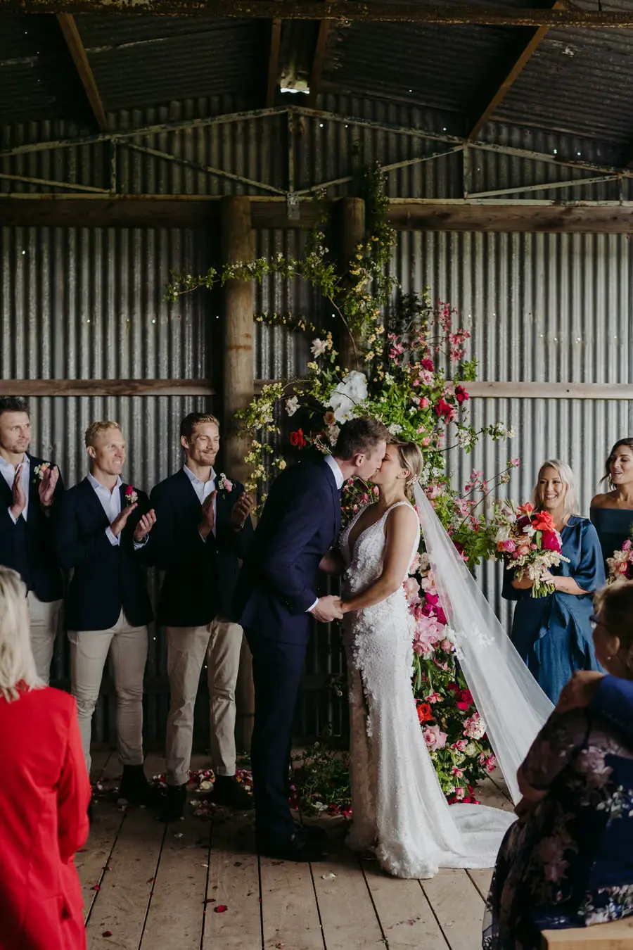 Bride and groom holding hands beneath a tall floral arrangement.