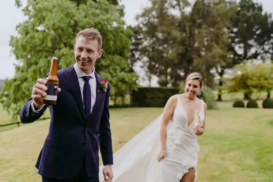 Groom raising a bottle while the bride follows across the lawn.