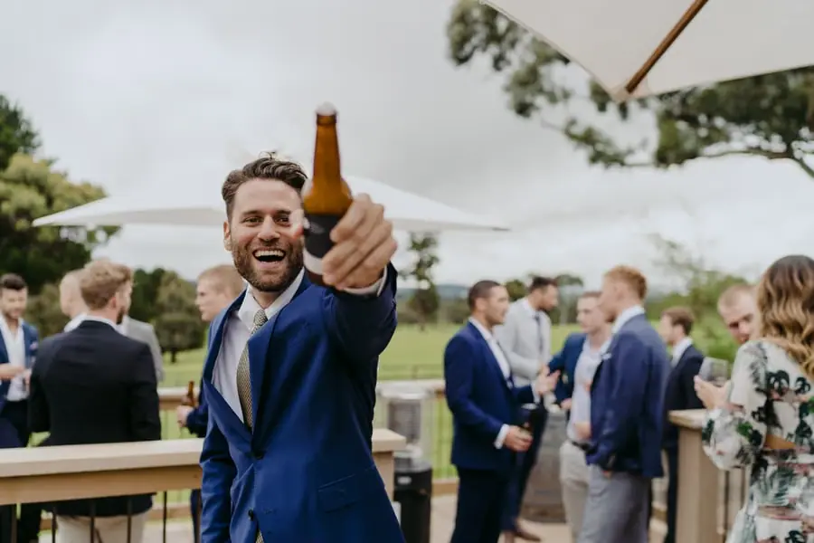 Man in a blue suit raising a bottle toward the camera outdoors.