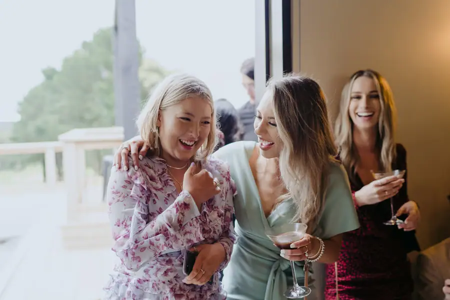 Three women laughing and holding drinks beside a reception window.