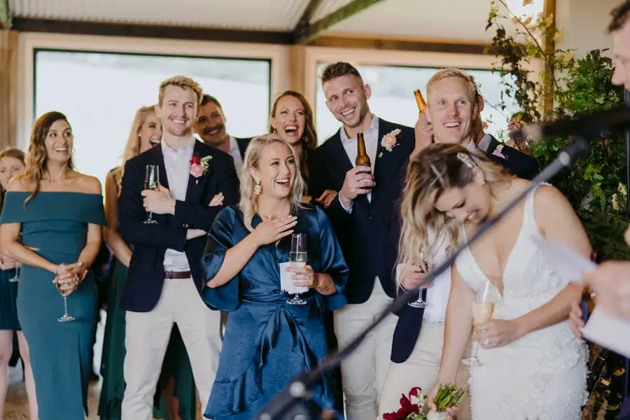 Bride leaning into an embrace as wedding party members laugh around her.
