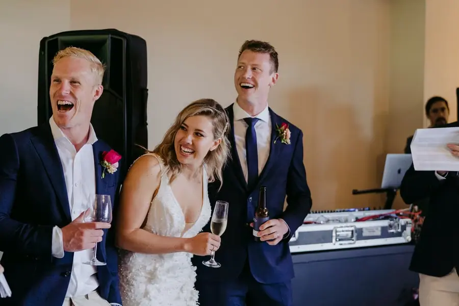 Bride and groom laughing beside a suited guest near the DJ table.