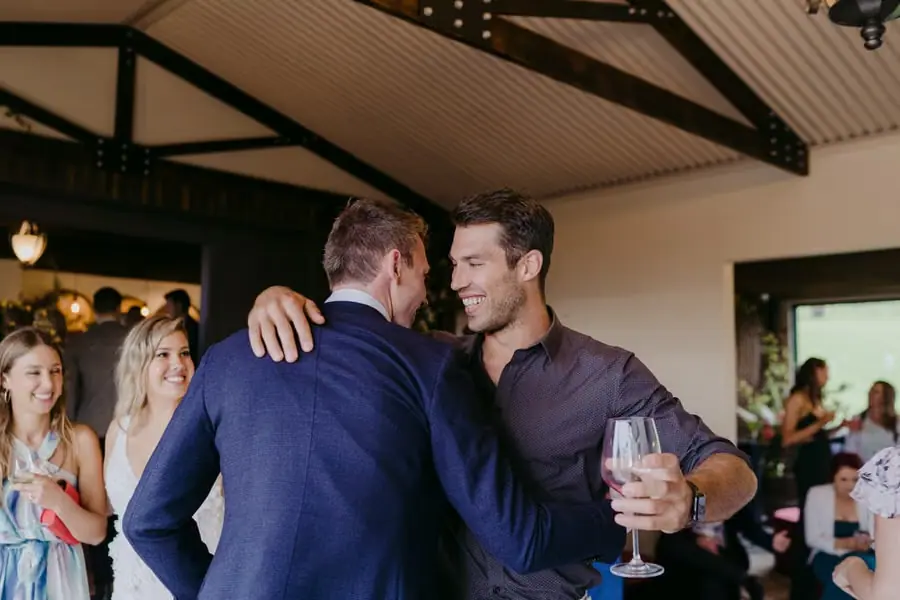 Groom embracing a man holding a wine glass in the reception room.