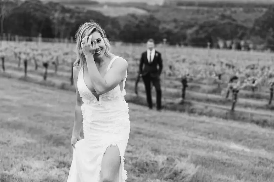 Black-and-white photograph of the bride smiling near vineyard rows with the groom behind her.