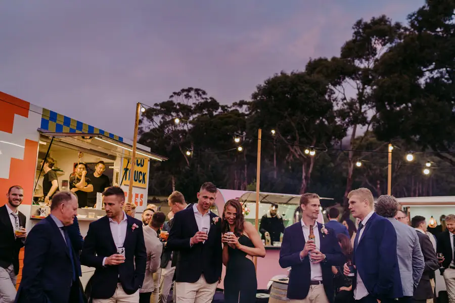 Guests chatting beside the taco truck under glowing string lights at dusk.