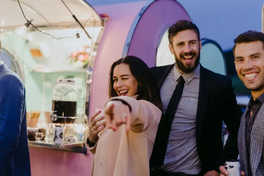 Three guests laughing beside a pink mobile bar at dusk.