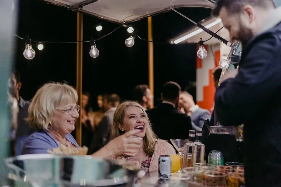Two women seated at an outdoor table laughing with a standing man.