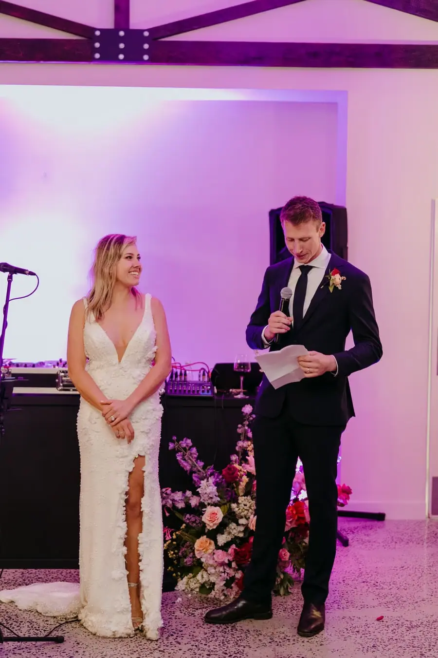 Groom reading notes into a microphone beside the bride under purple lighting.