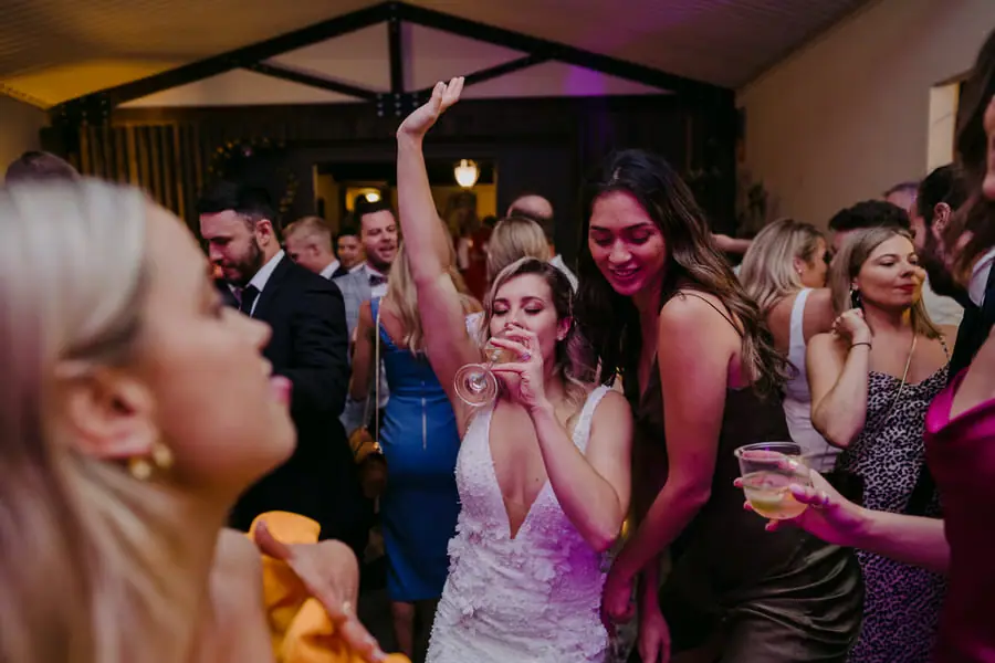 Bride dancing with women holding drinks beneath purple lights.