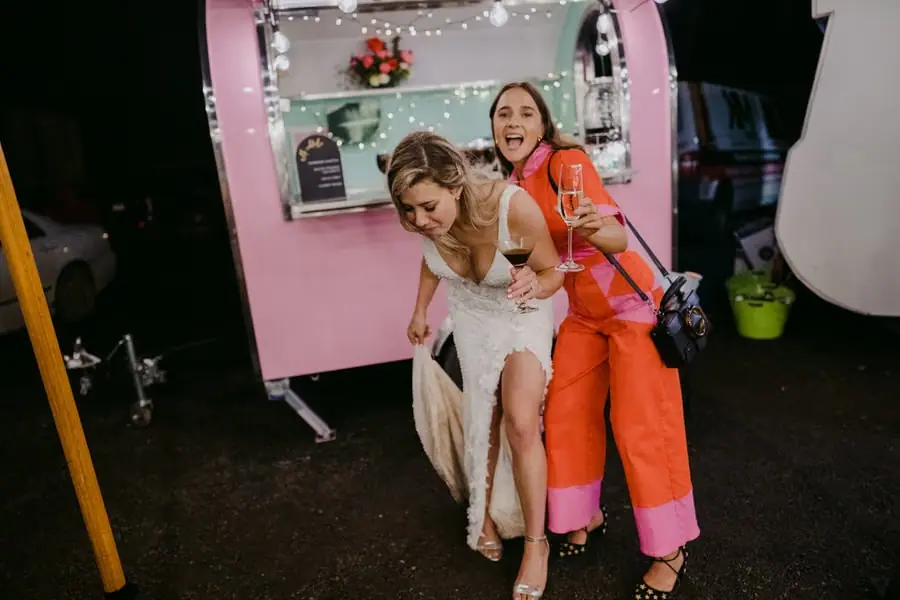 Woman in an orange outfit embracing the bride beside the pink mobile bar.