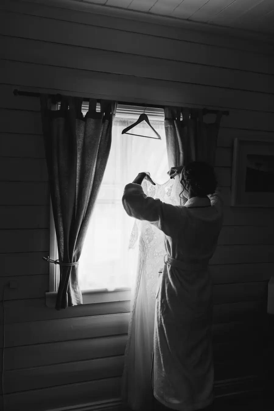 Black-and-white photograph of a woman reaching up to the wedding dress hanging in a window