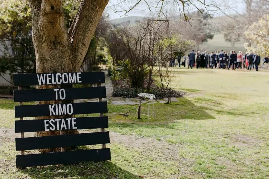 Welcome to Kimo Estate sign with wedding guests gathered beyond the trees