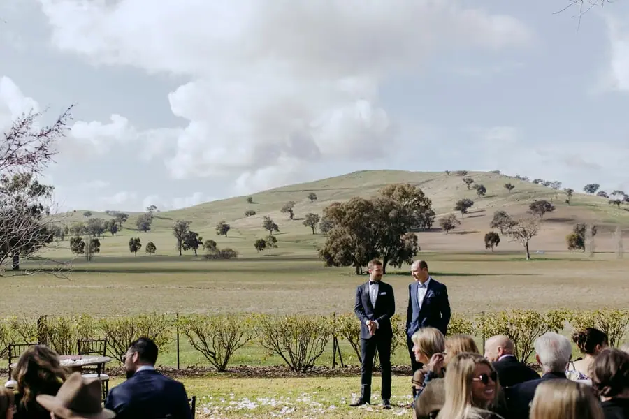 Groom and another man standing before seated wedding guests with hills behind them