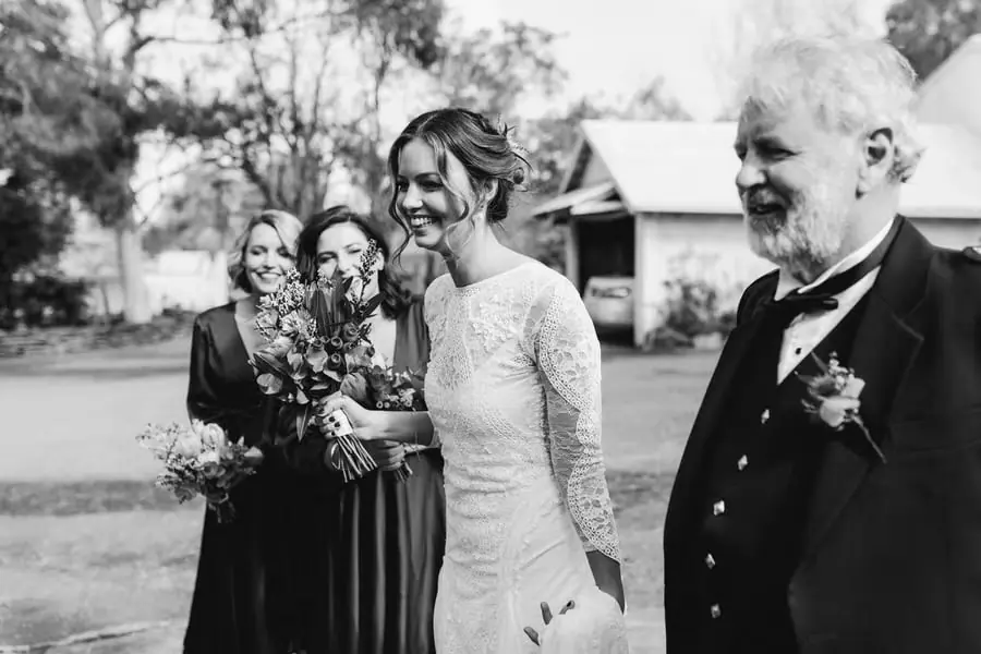 Black-and-white photograph of the bride walking with an older man as two bridesmaids follow