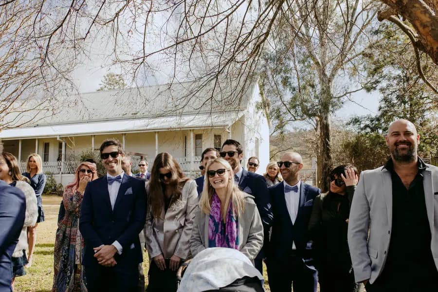 Wedding guests smiling beneath the trees outside a white cottage