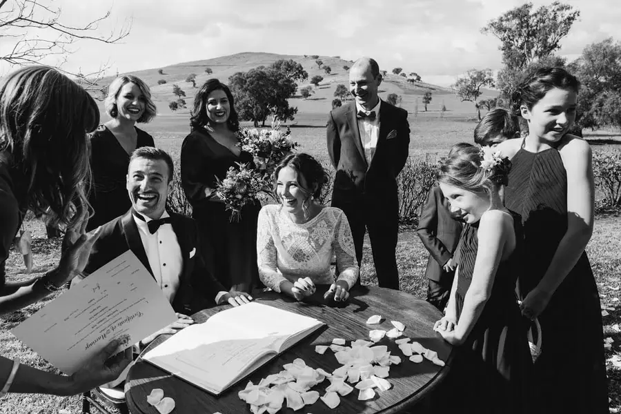Black-and-white photograph of the bride and groom laughing at the signing table surrounded by attendants