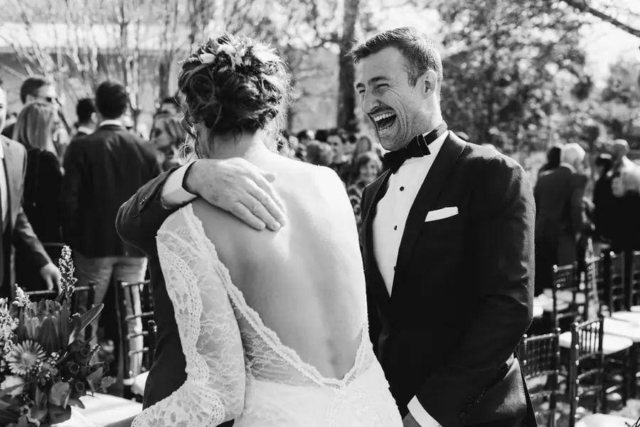 Black-and-white photograph of the groom laughing as he embraces the bride among guests