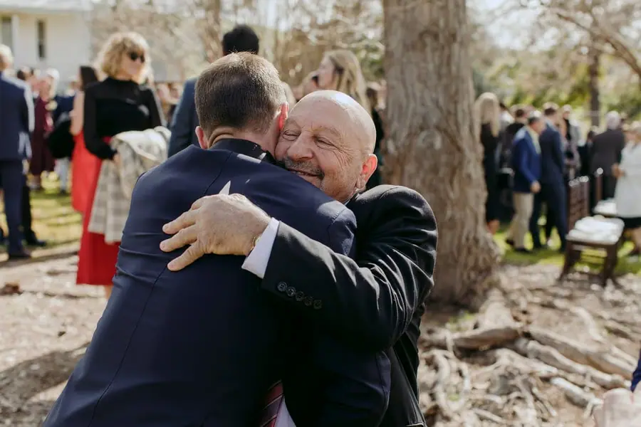 Groom embracing a smiling older man outside among wedding guests