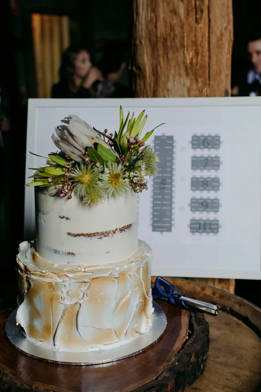 Two-tier wedding cake with flowers and gold decoration beside a seating chart