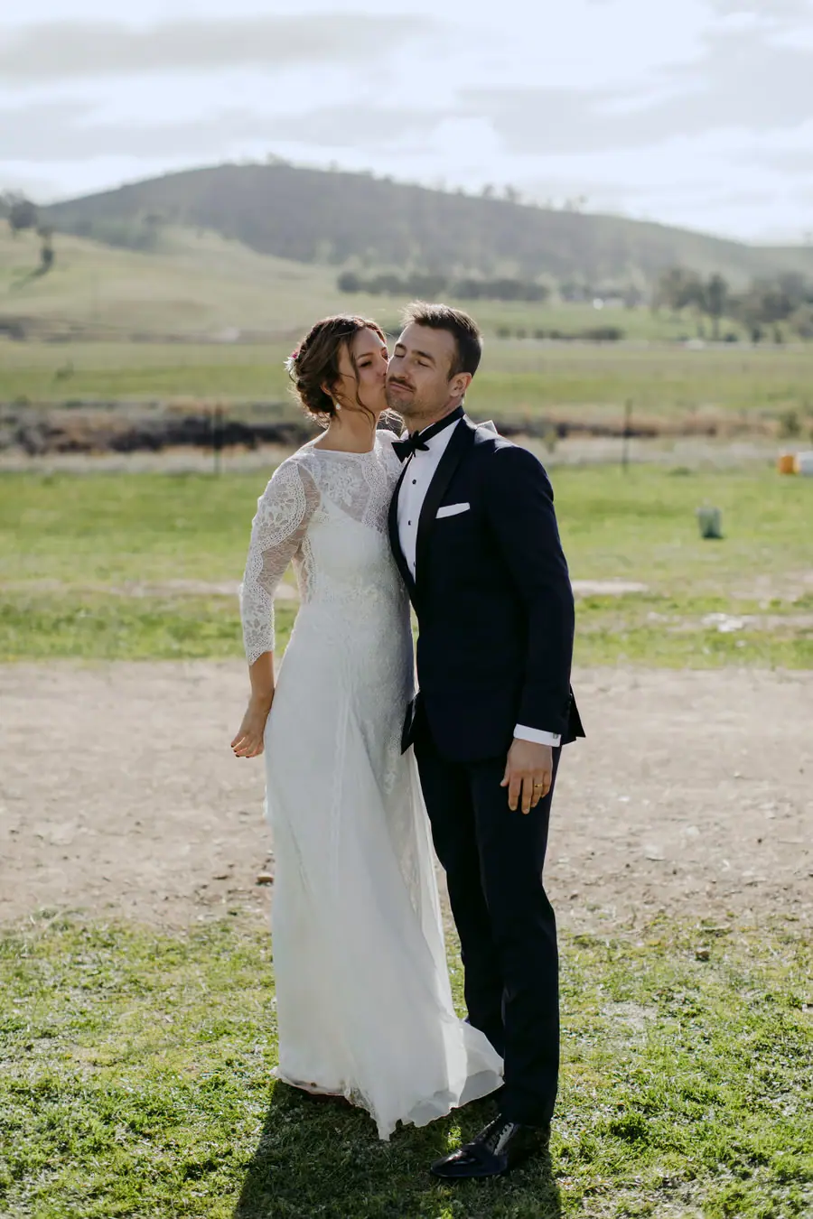 Bride kissing the groom’s cheek outside with farmland behind them