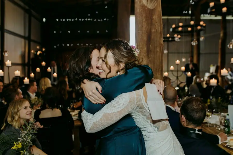 Bride embracing a woman in a teal dress among reception guests
