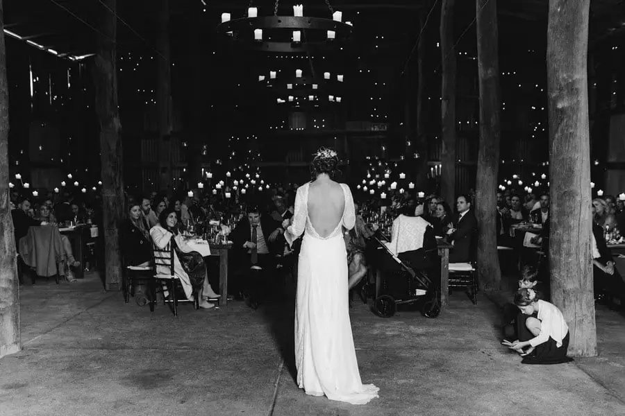 Black-and-white photograph of the bride standing between reception tables with her back to the camera