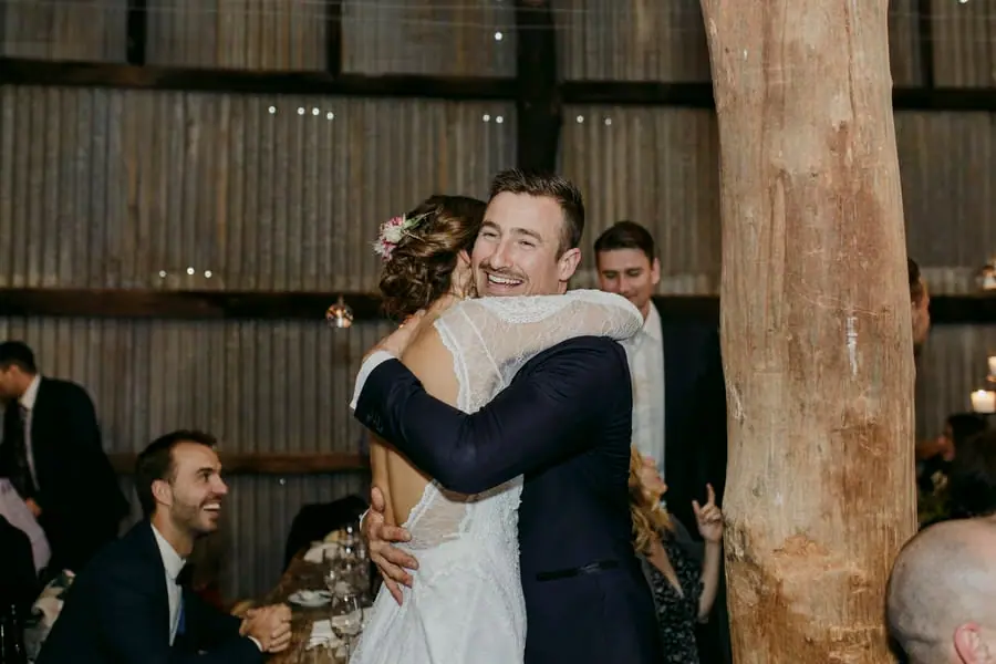 Bride and groom embracing beside a timber post as guests watch