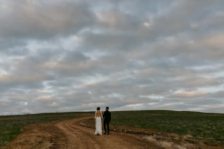 Bride and groom walking along a farm track beneath a broad cloudy sky