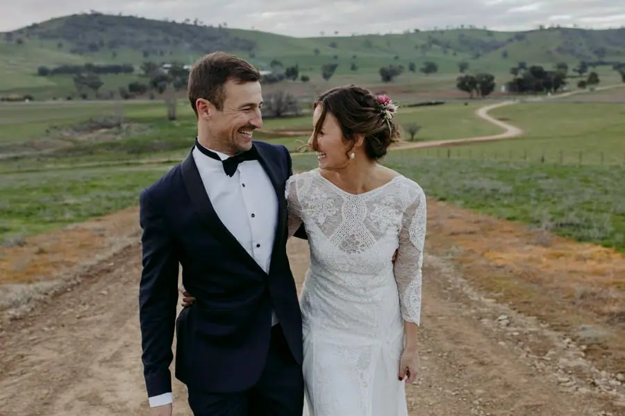 Bride and groom smiling at each other on a hillside above green farmland
