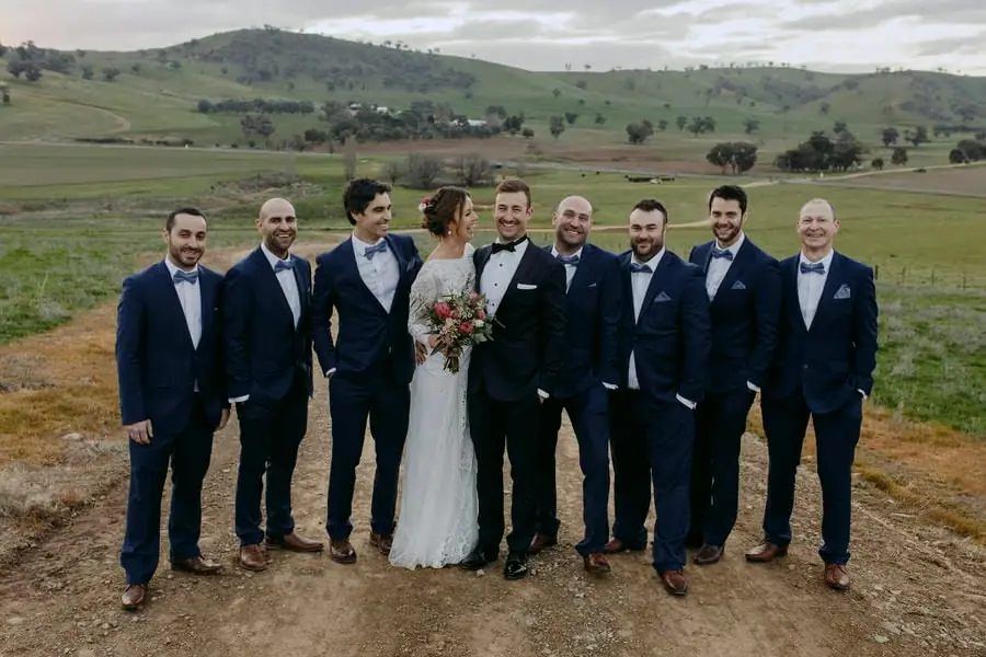 Bride and groom posing with a line of men in blue suits on a hillside