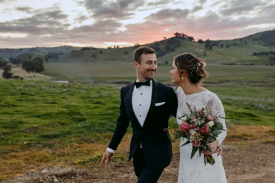 Bride and groom looking at each other beneath a pink sunset above green hills