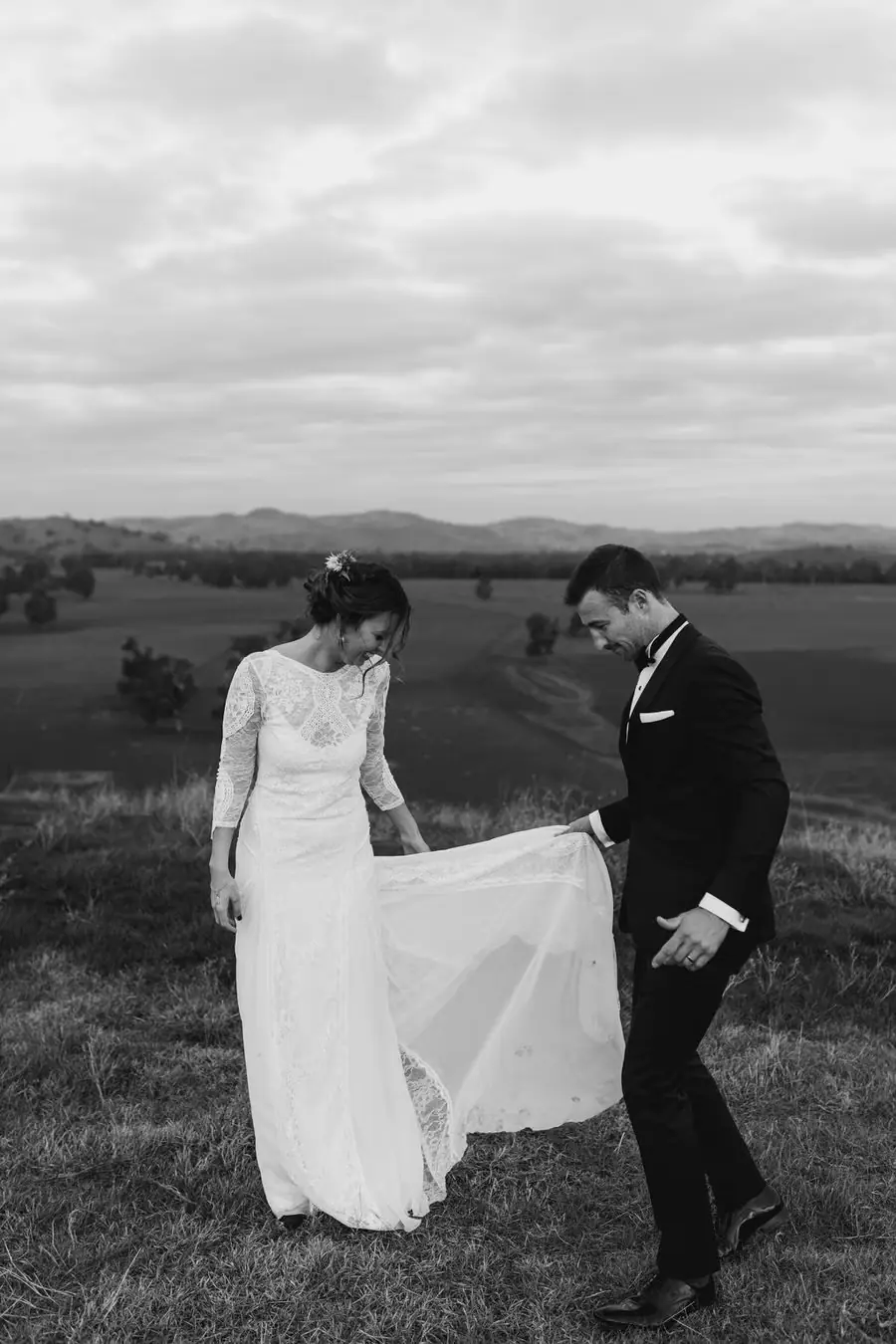 Black-and-white photograph of the groom lifting the edge of the bride’s dress on a hillside