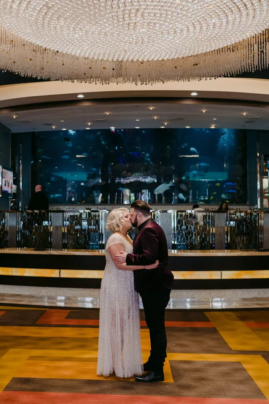 Bride and groom standing together beneath a large hotel-lobby chandelier