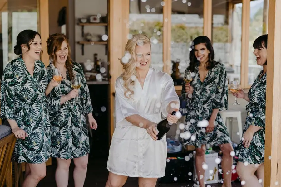 Bride opening a bottle of champagne as friends in matching robes laugh