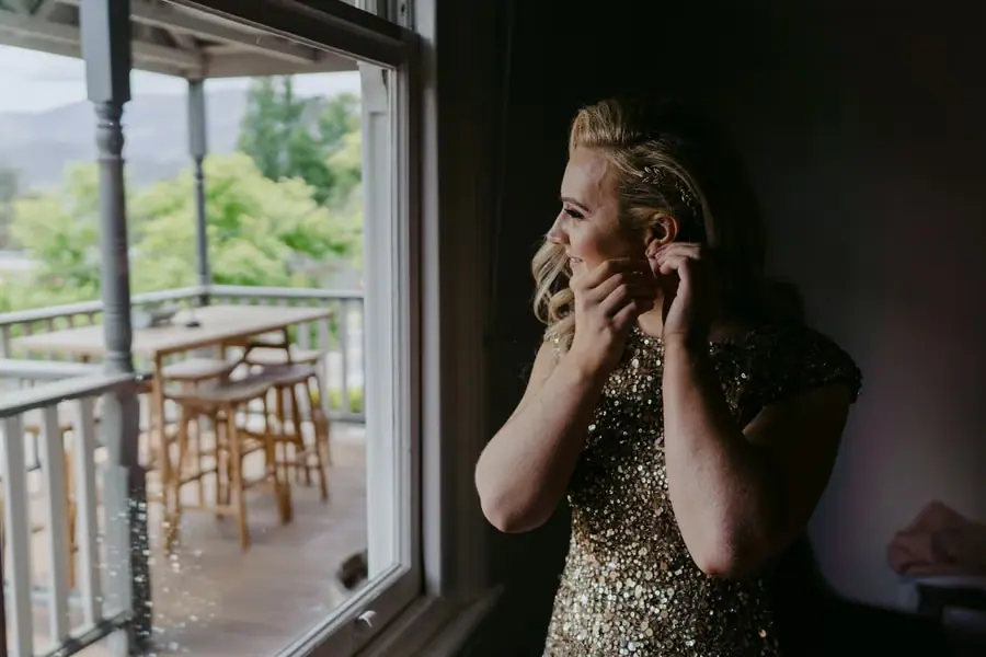 Bride fastening an earring beside a window while wearing a gold sequinned dress
