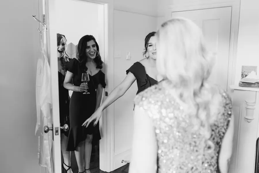 Black-and-white photograph of friends reacting as a bride enters in her gold dress