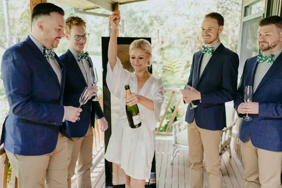 Bride raising an arm with a champagne bottle among four friends in blue jackets