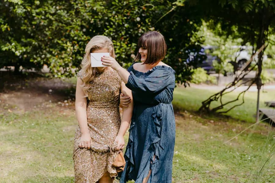 Friend holding a card over the eyes of a bride in a gold dress outdoors