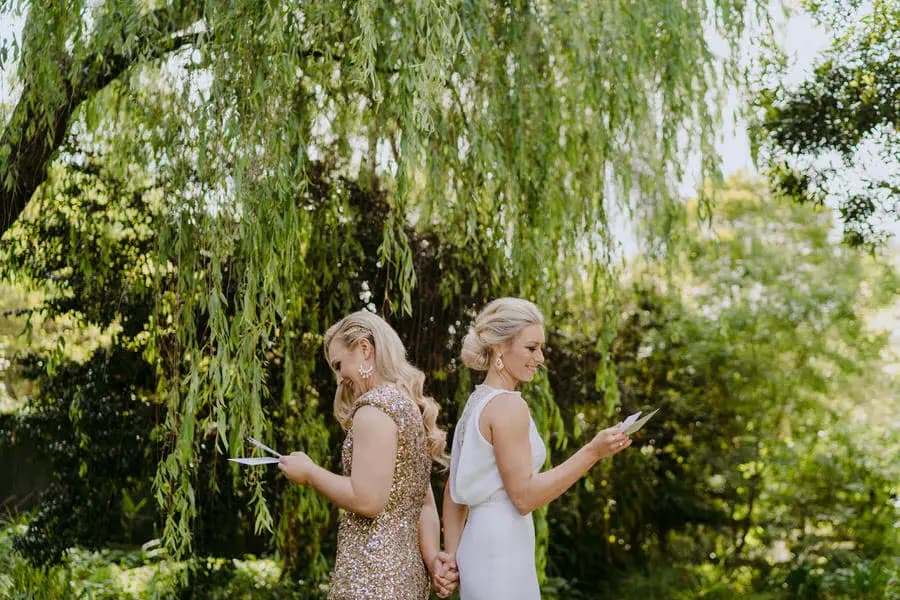 Two brides standing back to back beneath willow branches while reading cards