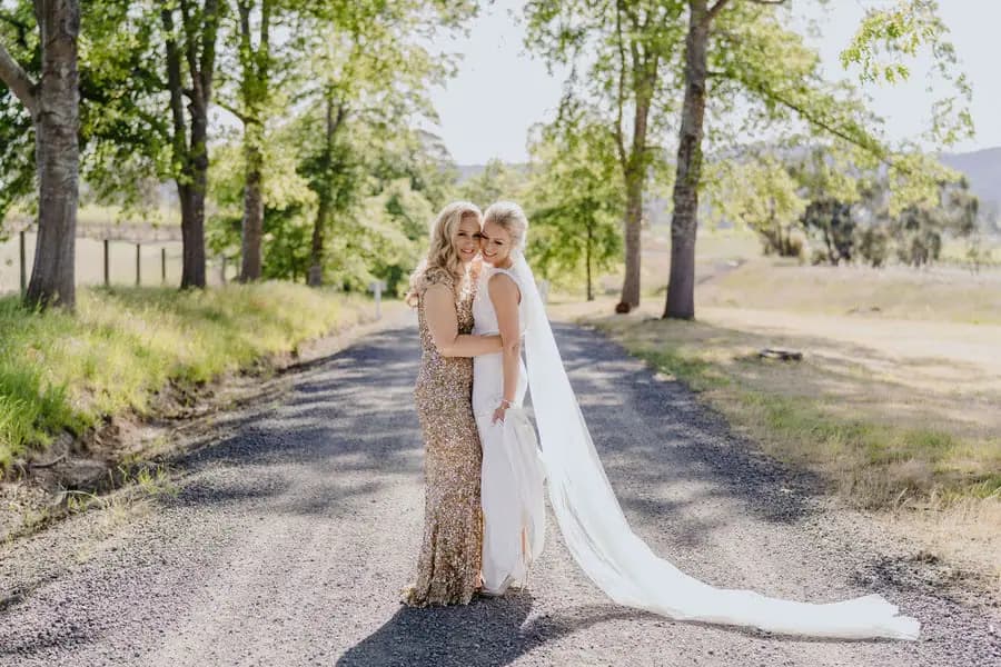 Two brides embracing on a tree-lined gravel driveway