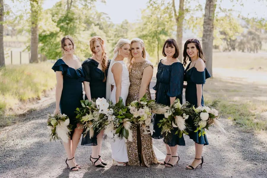 Two brides posing with four women in dark teal dresses and white bouquets