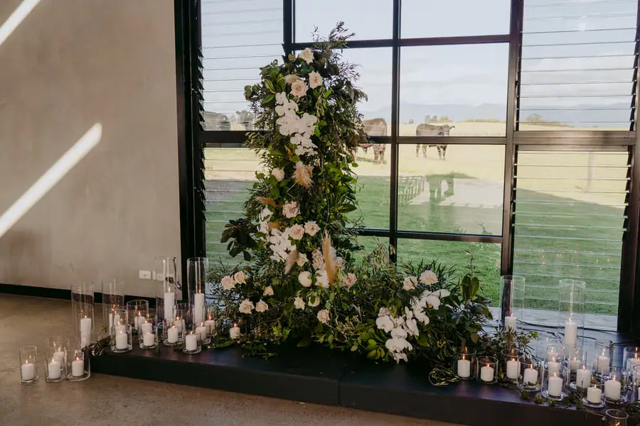 White floral arrangement and glass candle holders beside a tall ceremony window
