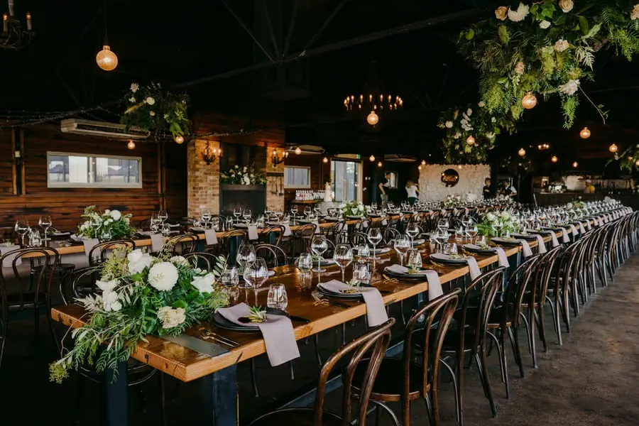 Long wooden reception tables set beneath warm lights and suspended greenery