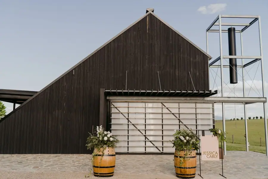 Dark timber ceremony building with flower-decorated barrels outside
