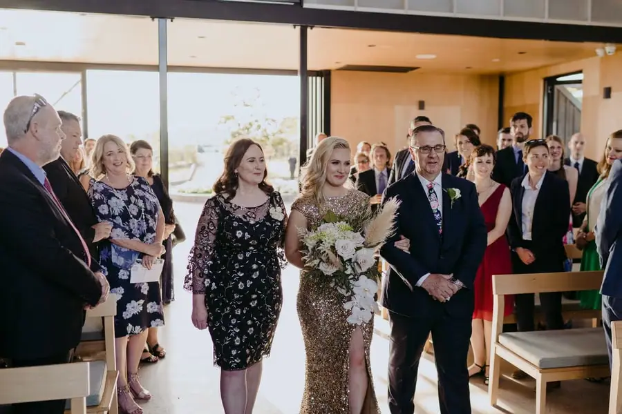Bride in a gold dress walking down the ceremony aisle beside two companions