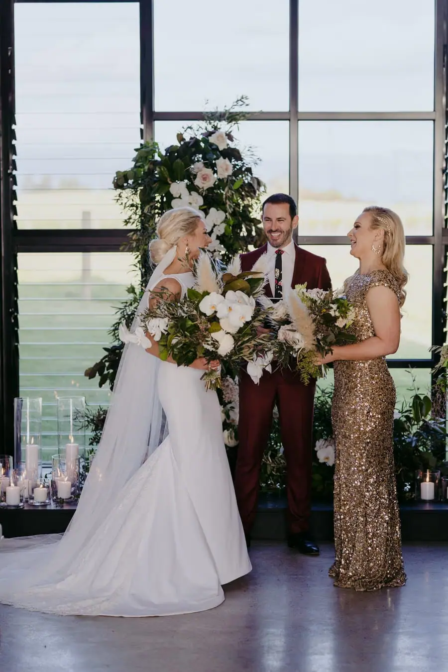 Two brides facing one another beside the celebrant and a tall floral arrangement