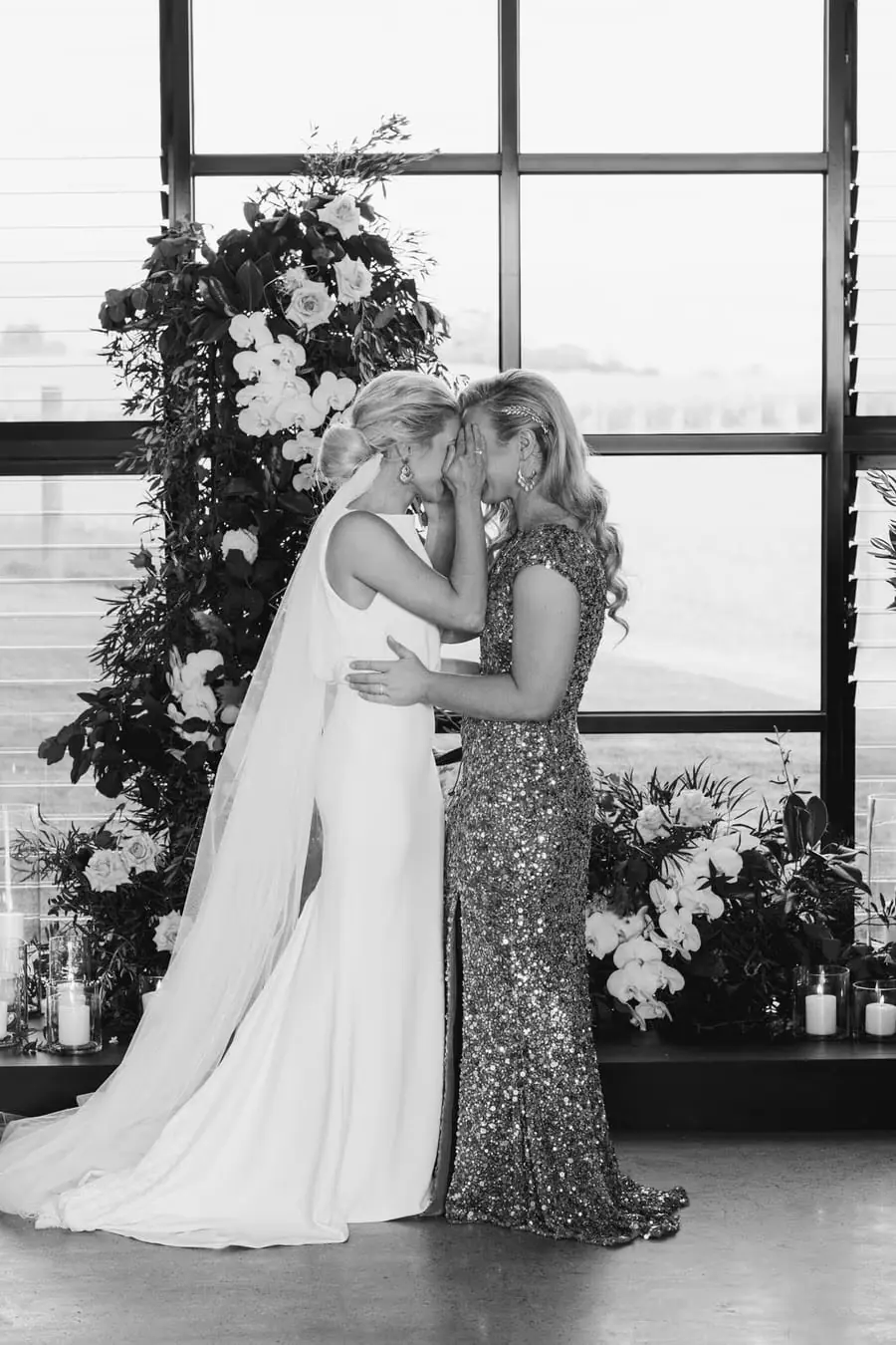 Black-and-white photograph of two brides kissing beside a floral ceremony arrangement
