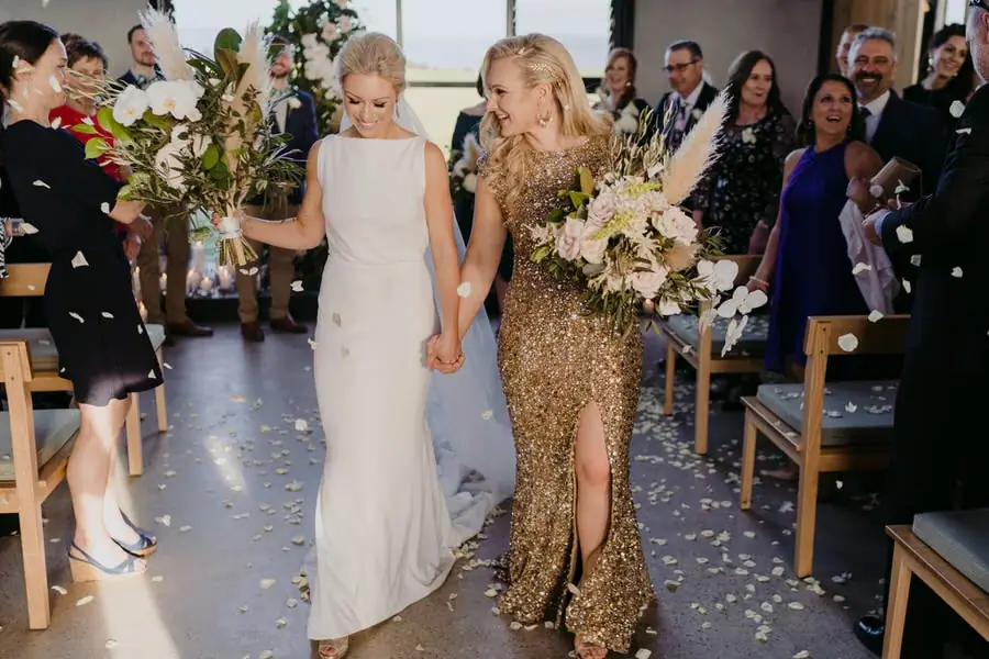 Two brides holding hands and walking down the aisle through falling confetti
